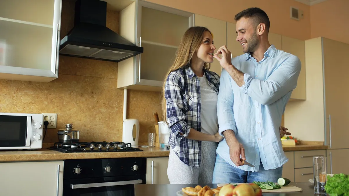 Paar kocht gemeinsam in einer Küche, der Mann hält der Frau lachend einen Bissen hin