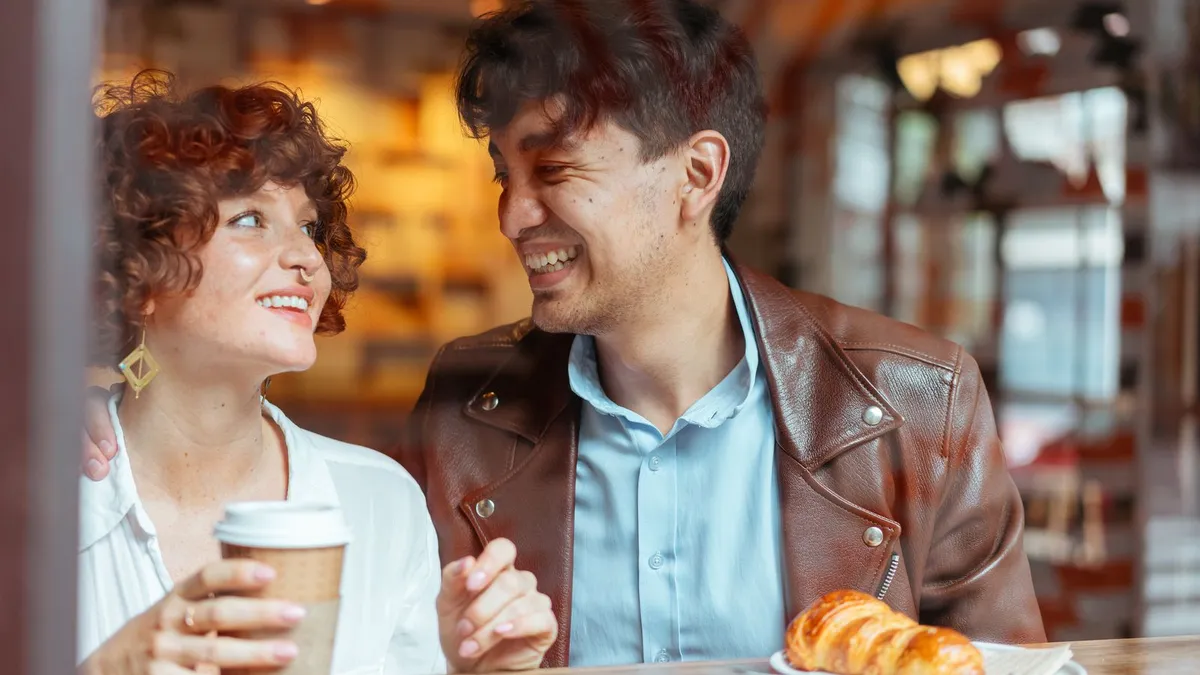 Lachendes Paar sitzt im Café nah beieinander, vor ihnen ein Croissant und ein Kaffeebecher