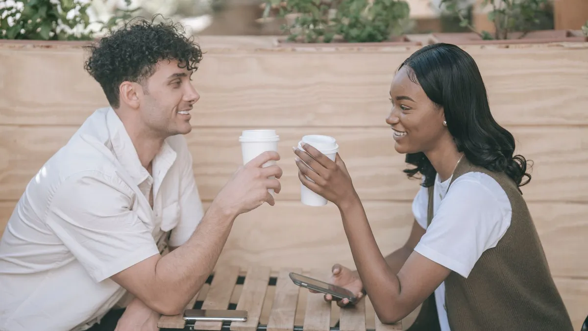Mann und Frau stoßen draußen mit Kaffeebechern an und lächeln sich an, auf dem Tisch liegt ein Smartphone