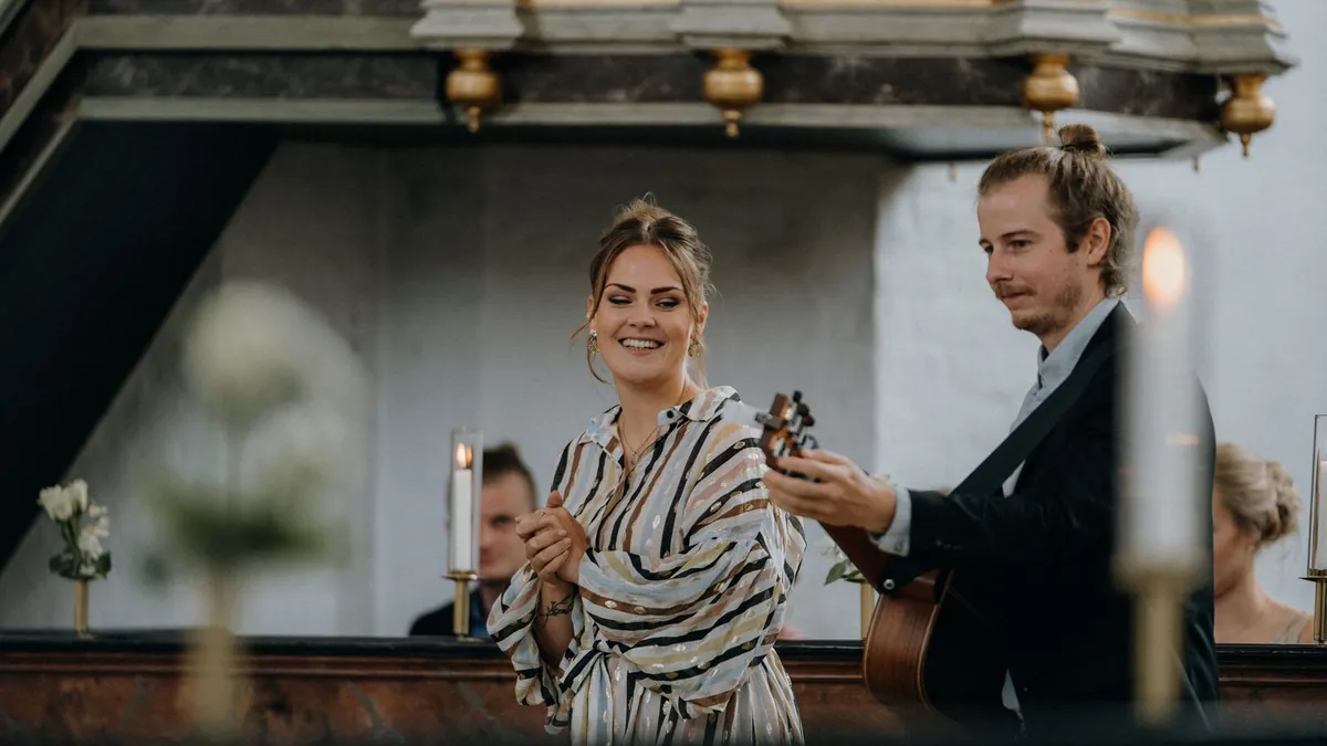 Eine Sängerin und ein Gitarrist musizieren lächelnd in einer Kirche, im Vordergrund unscharfe Kerzen