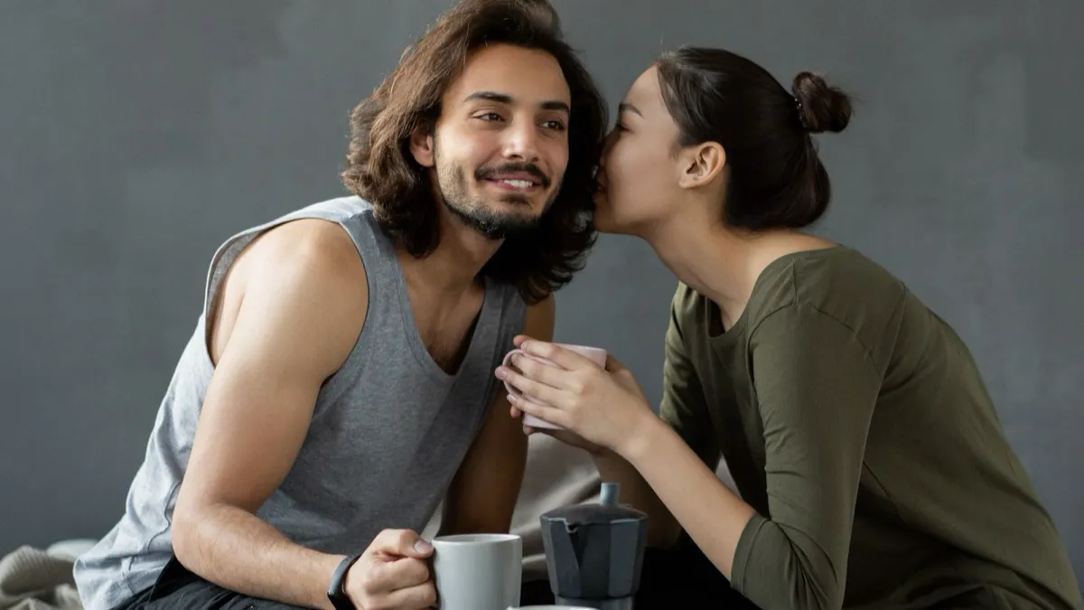 Junges Paar sitzt mit Kaffeetassen auf einem Bett aus Holzpaletten, sie flüstert ihm etwas ins Ohr