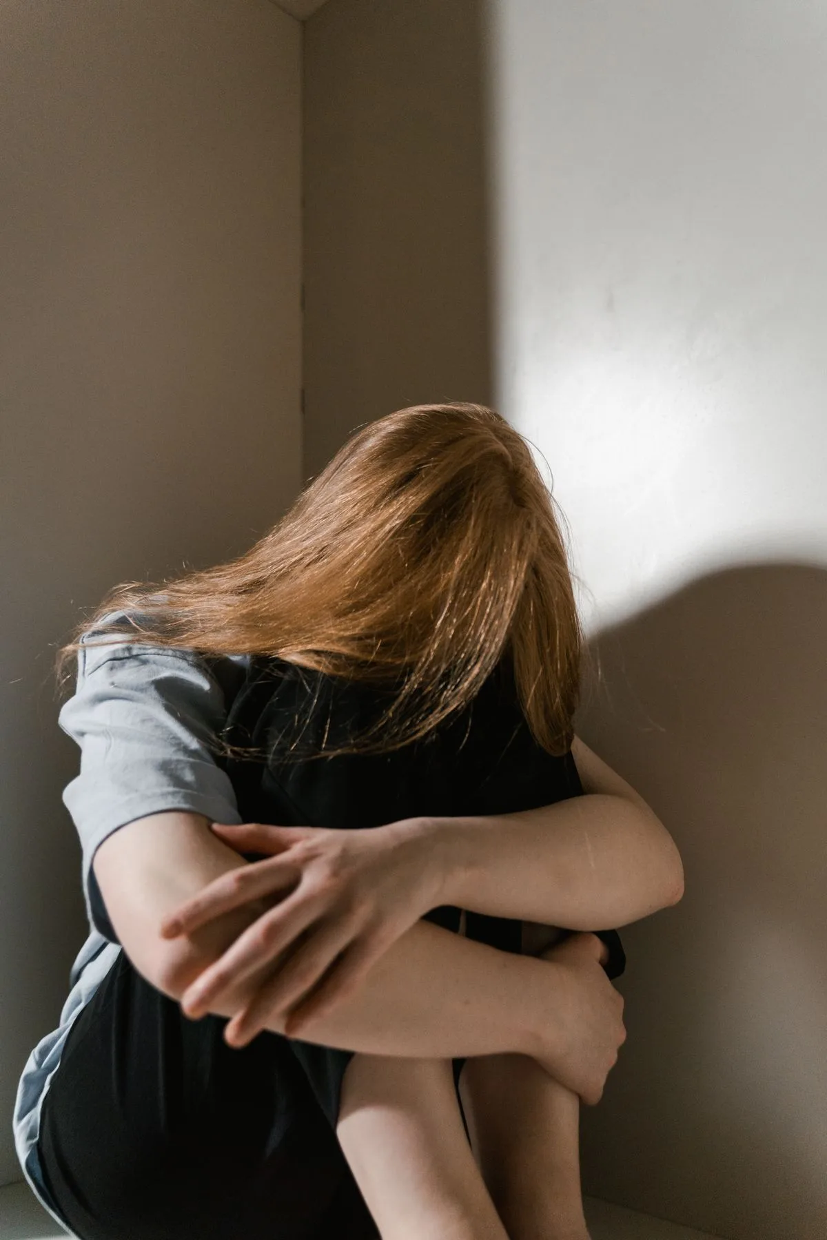 Frau sitzt zusammengekauert in einer Zimmerecke, das Gesicht hinter den Haaren verborgen