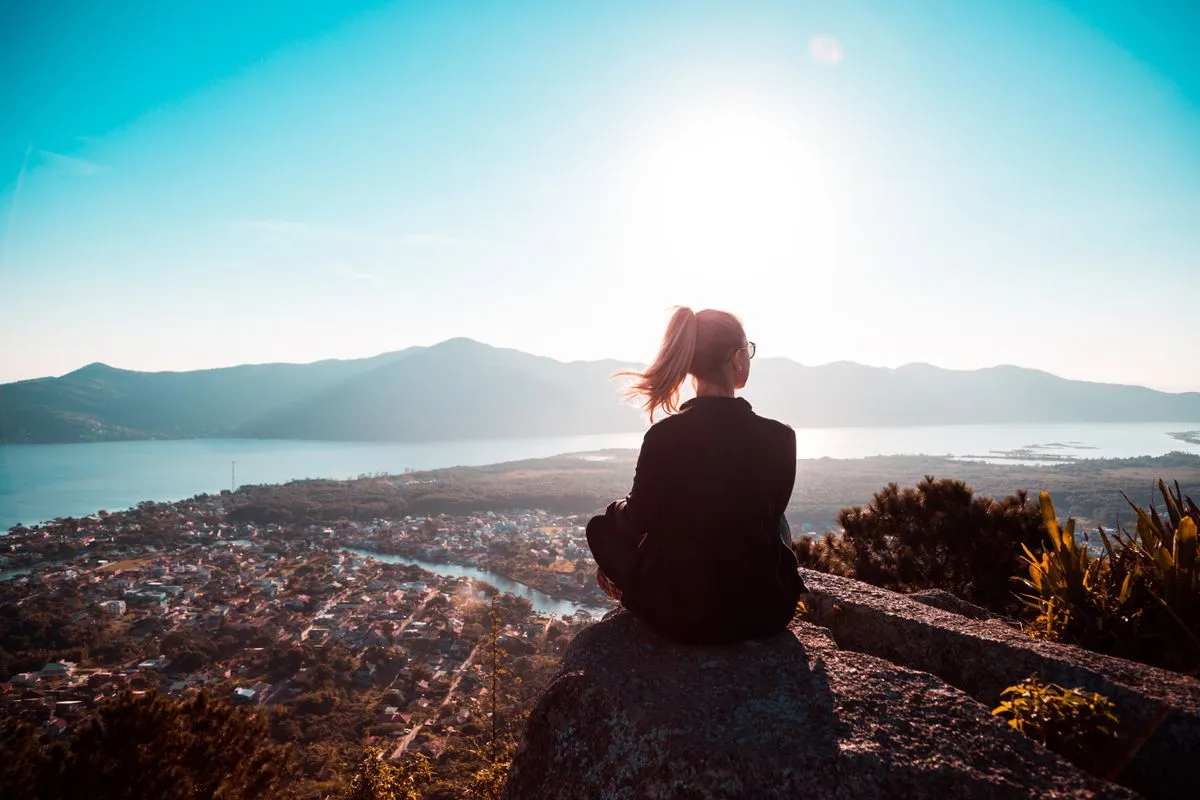 Frau sitzt ruhig auf einem Felsen und blickt bei Sonnenlicht über eine weite Landschaft mit See
