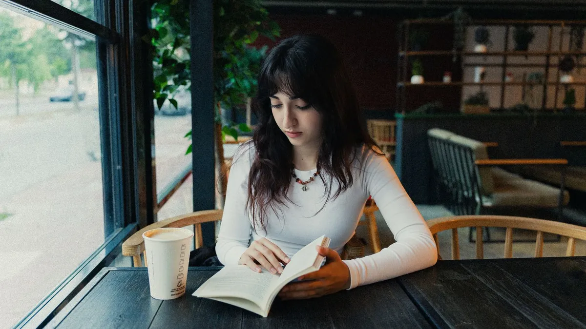 Junge Frau liest in einem ruhigen Café ein Buch, neben ihr steht ein Kaffeebecher