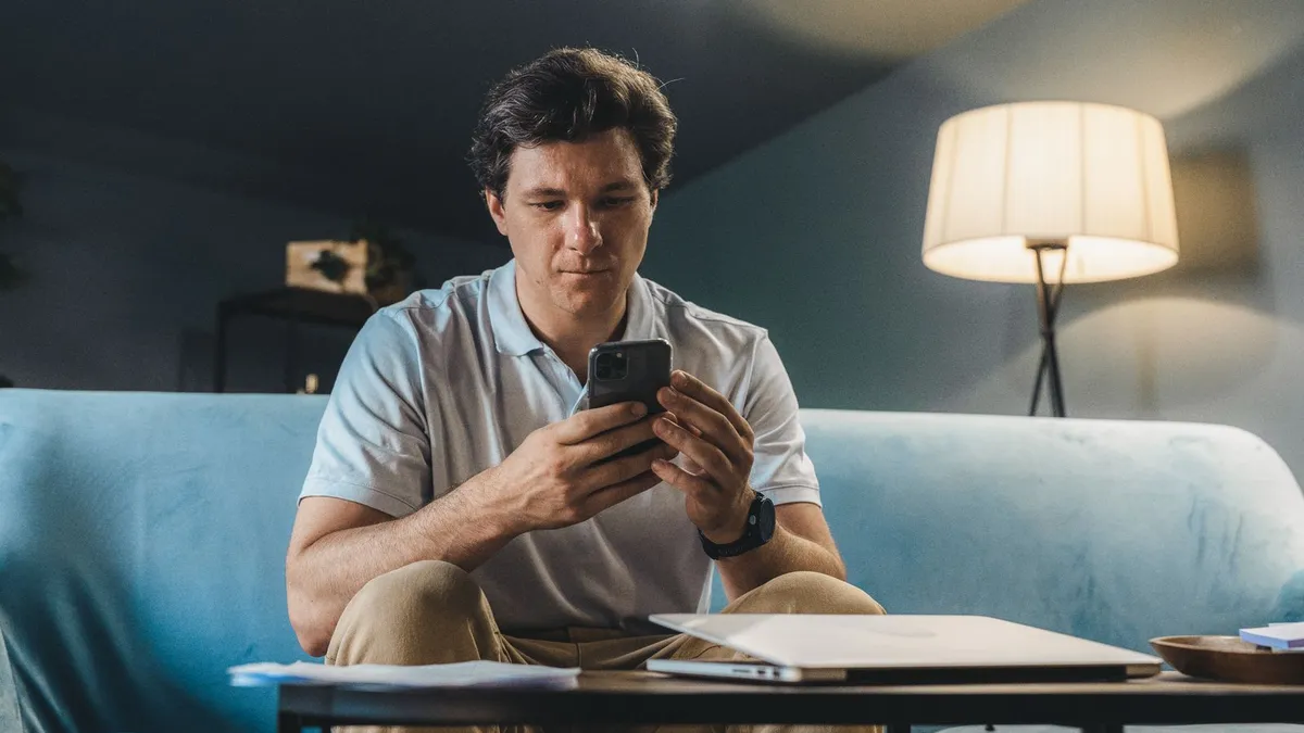 Mann sitzt abends auf dem Sofa und schaut auf sein Smartphone, daneben eine Stehlampe