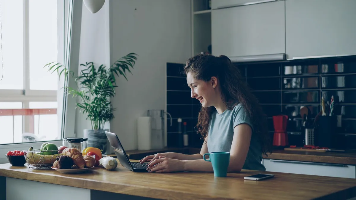Lachende junge Frau arbeitet in einer hellen Küche am Laptop, daneben stehen Obstschale und Tasse