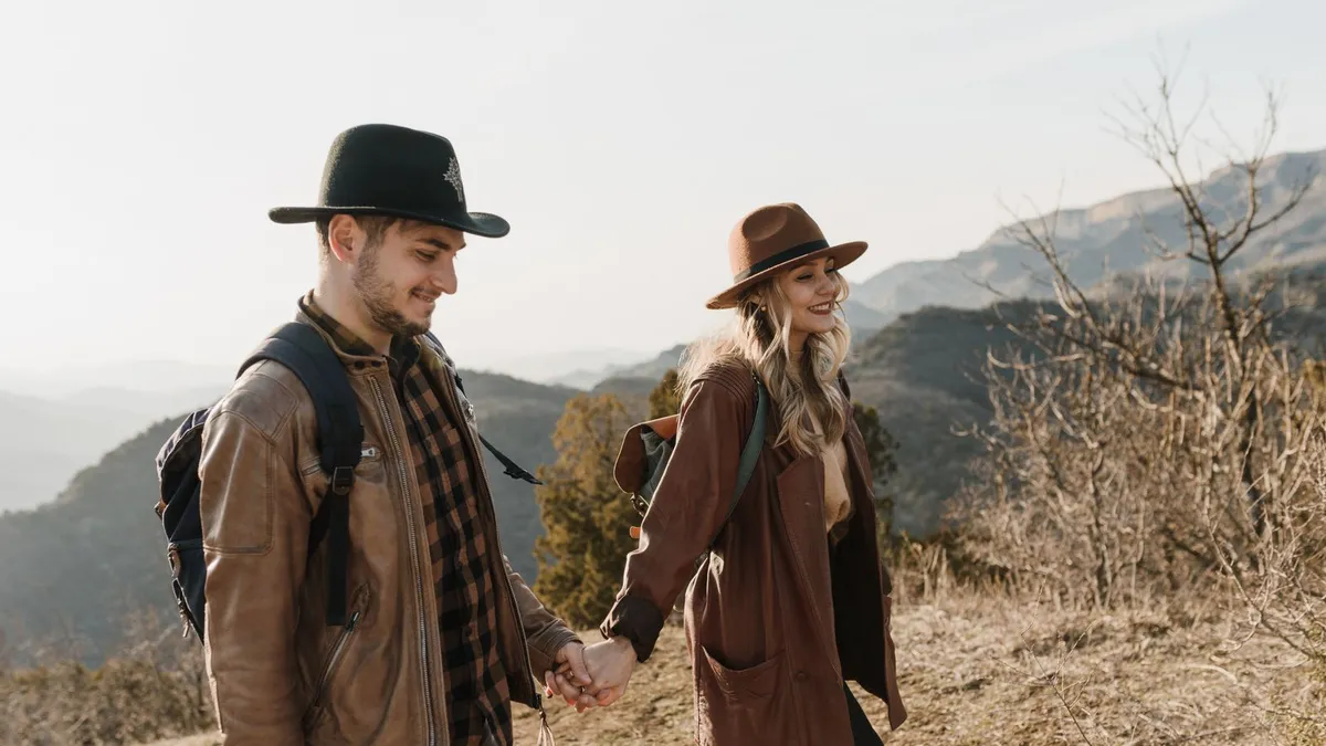 Lächelndes Paar mit Hüten und Rucksäcken wandert Hand in Hand durch eine Berglandschaft