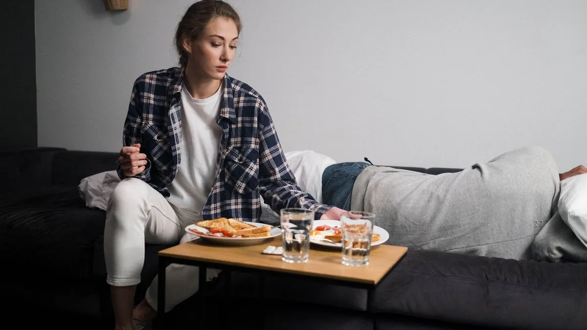 Frau in kariertem Hemd sitzt vor einem gedeckten Frühstückstisch, hinter ihr liegt ein Mann auf dem Sofa