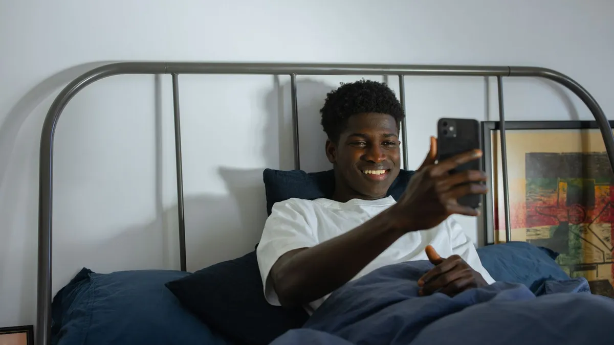 Ein junger Mann liegt im Bett und lächelt beim Blick auf sein Smartphone