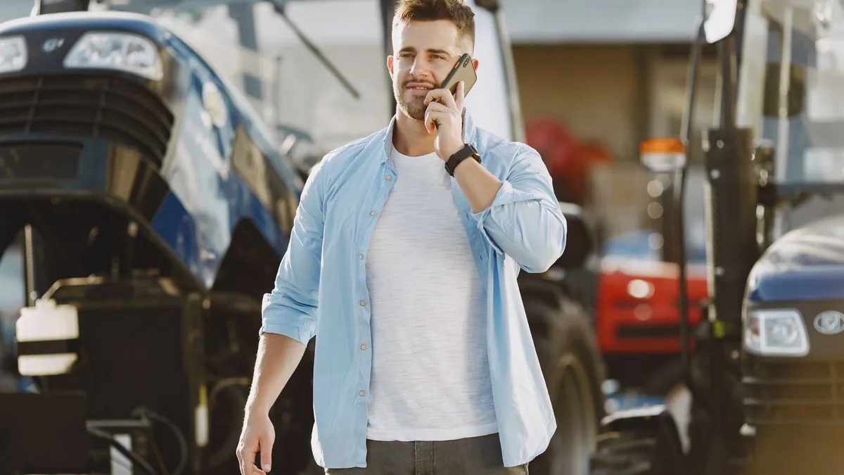 Junger Mann telefoniert mit dem Handy, hinter ihm stehen Traktoren