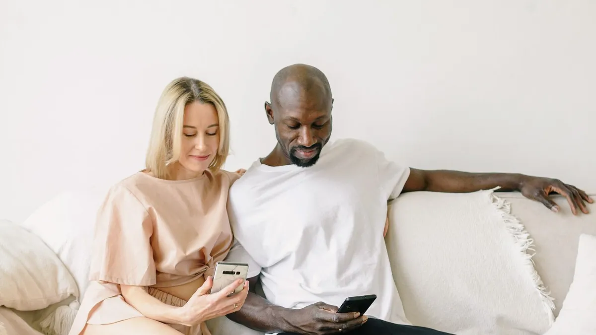 Paar sitzt nebeneinander auf einem hellen Sofa, beide schauen auf ihre Smartphones