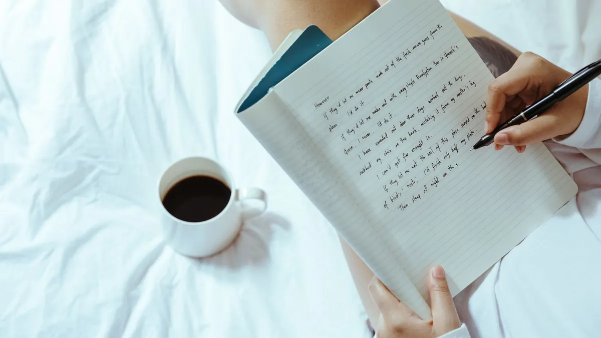 Hand schreibt mit einem Stift in ein Notizbuch, daneben steht eine Tasse Kaffee auf weißer Bettwäsche