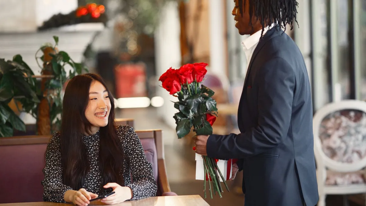 Lächelnde Frau sitzt in einem Café, neben ihr steht ein Mann im Anzug mit einem großen Strauß roter Rosen