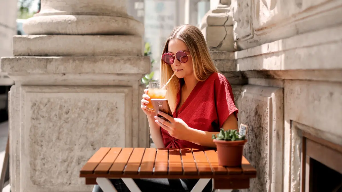 Junge Frau mit Sonnenbrille trinkt auf einer Caféterrasse und schaut auf ihr Smartphone