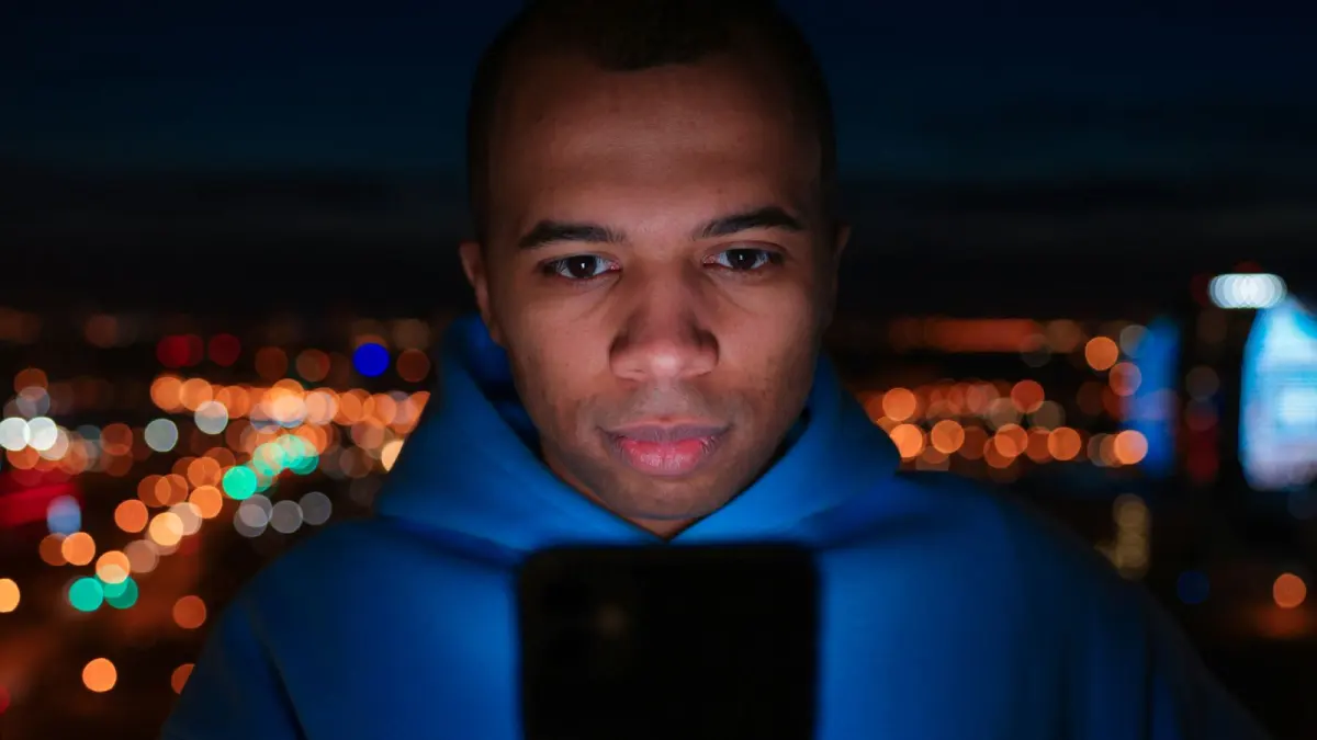 Junger Mann im blauen Hoodie schaut abends vor Stadtlichtern auf sein Smartphone
