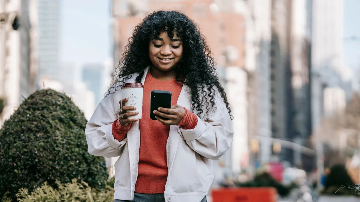 Lächelnde Frau mit lockigem Haar geht mit einem Kaffeebecher durch eine Stadtstraße und schaut auf ihr Smartphone