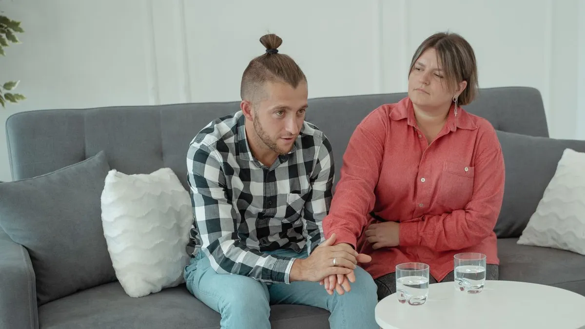 Ein Paar sitzt nebeneinander auf einem grauen Sofa und hält sich an den Händen, die Frau blickt ernst zur Seite