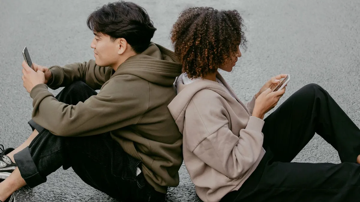 Zwei Menschen sitzen Rücken an Rücken auf dem Asphalt und schauen jeder auf sein eigenes Smartphone