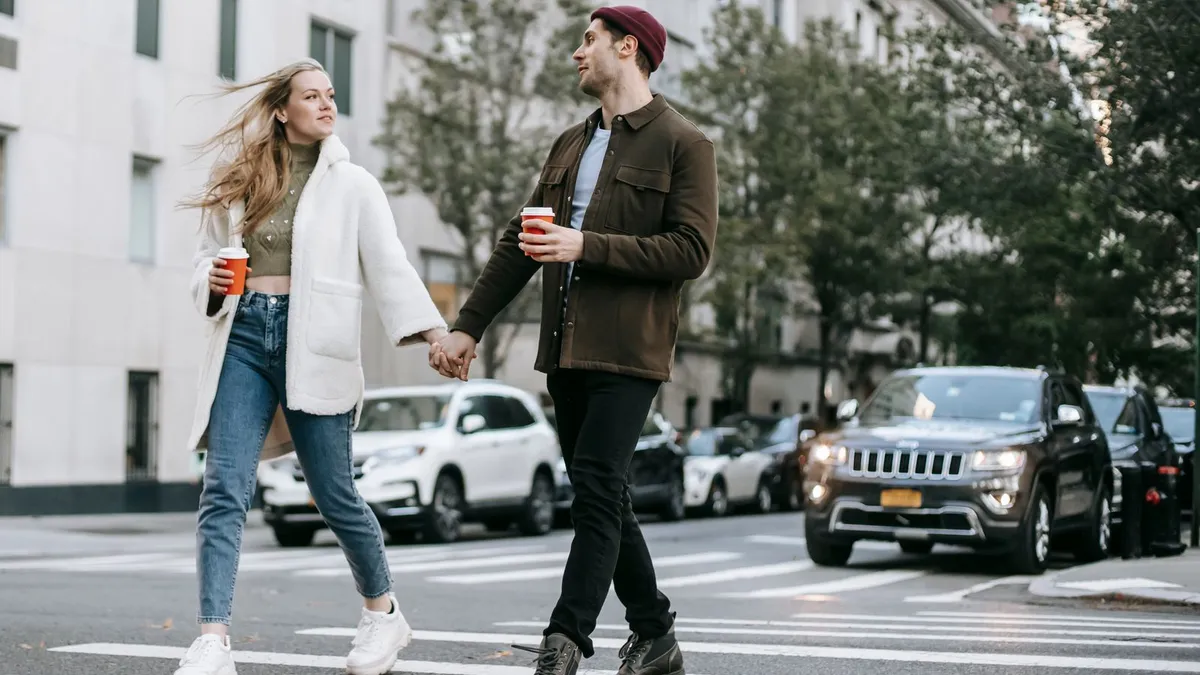 Junges Paar überquert Hand in Hand eine Straße, beide tragen Kaffeebecher zum Mitnehmen