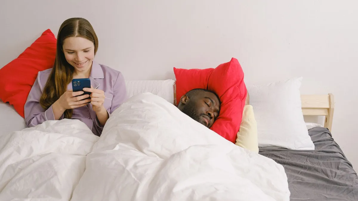 Frau lächelt im Bett auf ihr Smartphone, neben ihr schläft ihr Partner