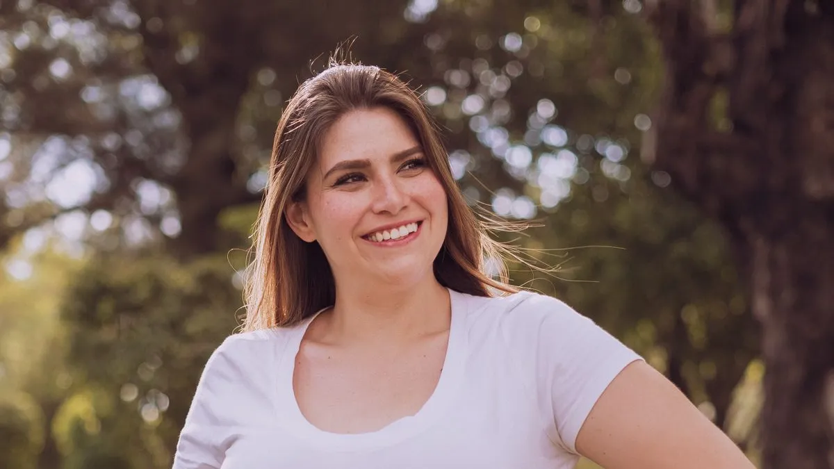Eine lächelnde Frau mit langen Haaren und weißem T-Shirt in einem sonnigen Park