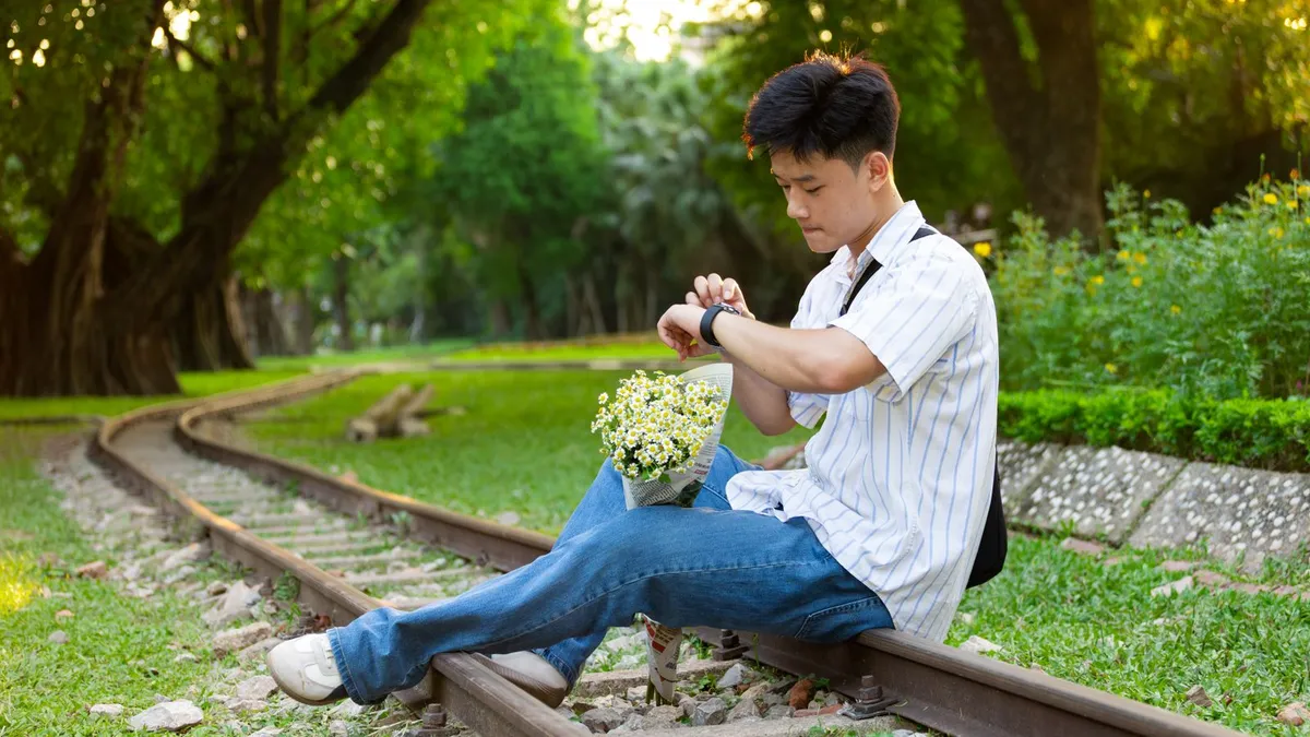 Junger Mann sitzt mit einem Blumenstrauß auf alten Bahngleisen in einem Park und schaut auf seine Armbanduhr