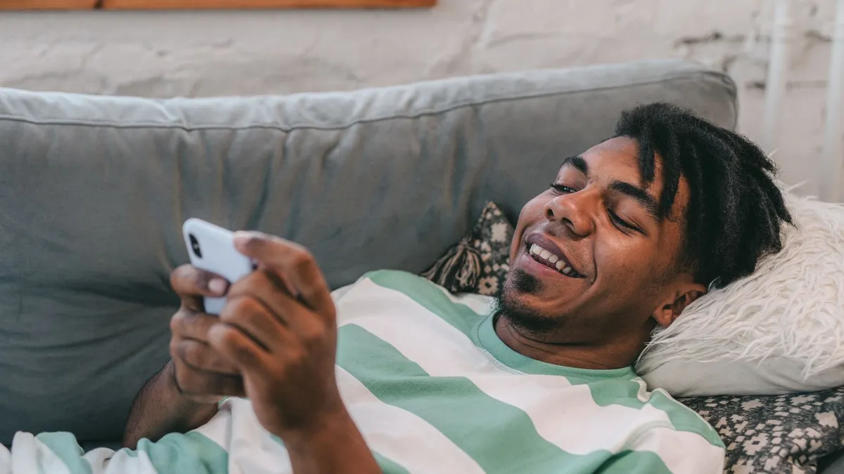 Lachender Mann liegt auf einem grauen Sofa und tippt auf seinem Smartphone