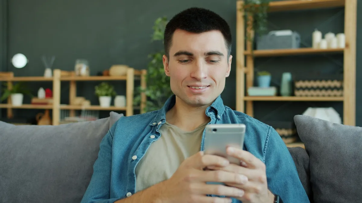Lächelnder Mann im Jeanshemd sitzt auf einem grauen Sofa und schaut auf sein Smartphone
