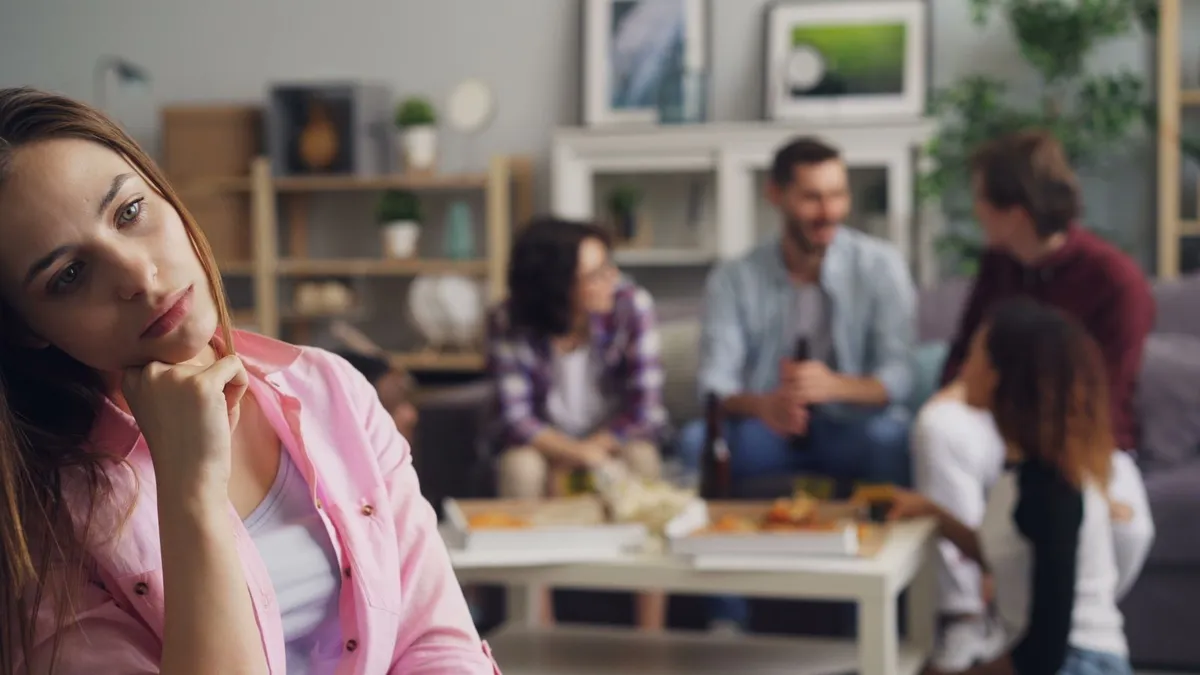 Frau mit Hand am Kinn schaut nachdenklich zur Seite, hinter ihr unterhält sich eine Gruppe am Couchtisch