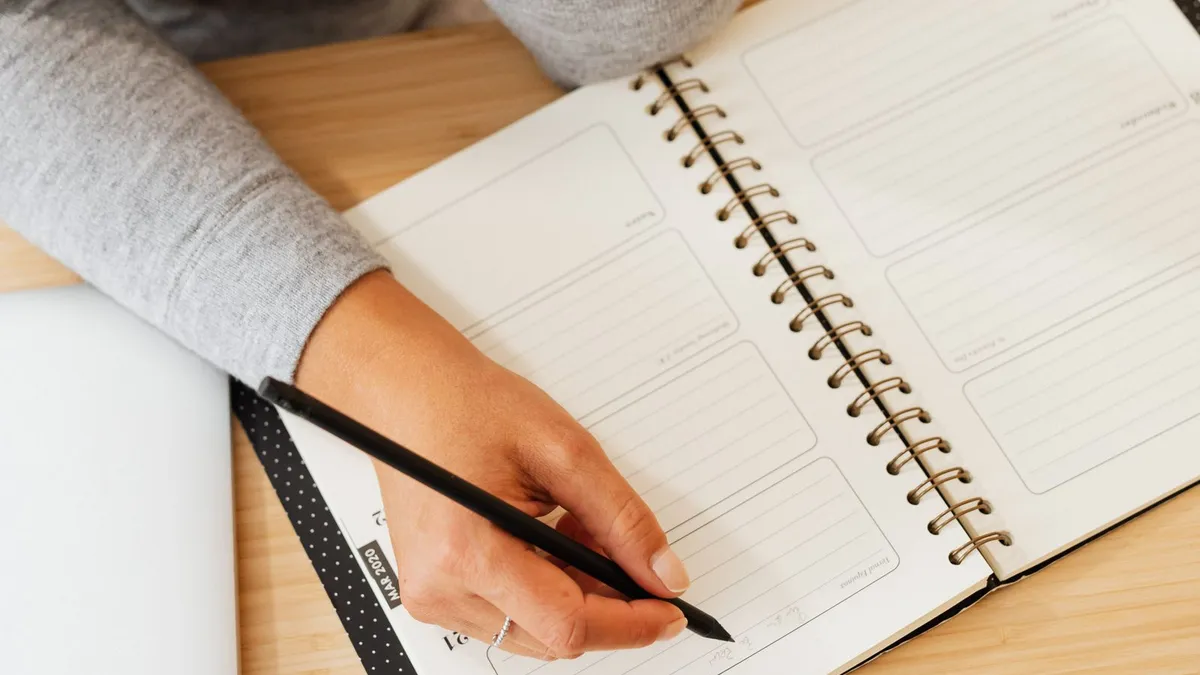 Hand mit Bleistift schreibt in einen aufgeschlagenen Wochenplaner mit Spiralbindung auf einem Holztisch