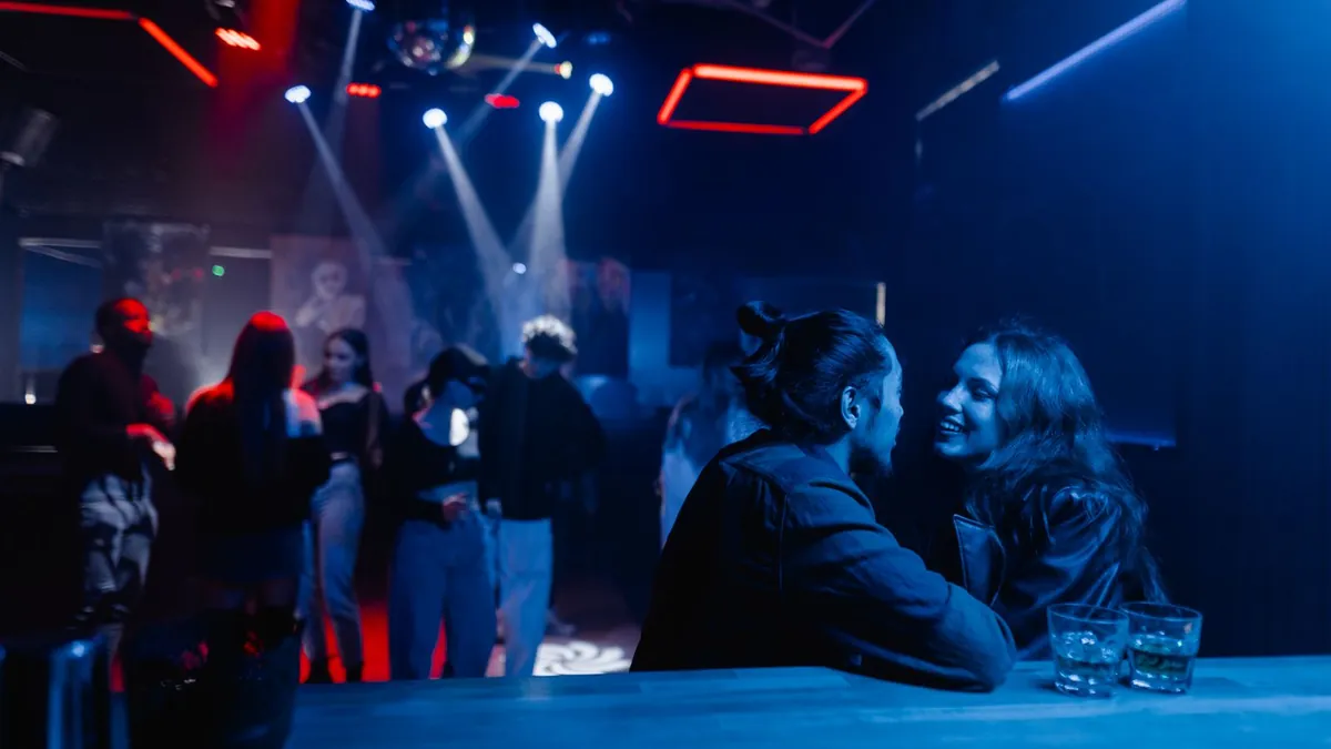 Ein junges Paar unterhält sich lächelnd an der Bar eines Clubs, im Hintergrund tanzen Menschen im blauen Licht