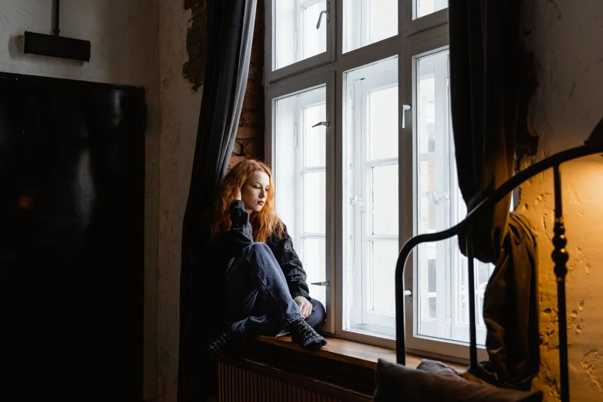 Nachdenkliche Frau sitzt still am Fenster, im unscharfen Hintergrund Spielzeug eines Kindes