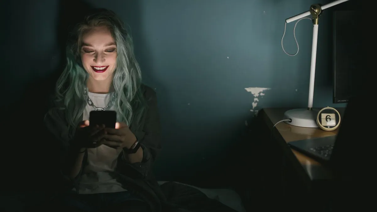 Frau mit blaugrünen Haaren lächelt in einem dunklen Zimmer auf ihr Smartphone, dessen Licht ihr Gesicht beleuchtet