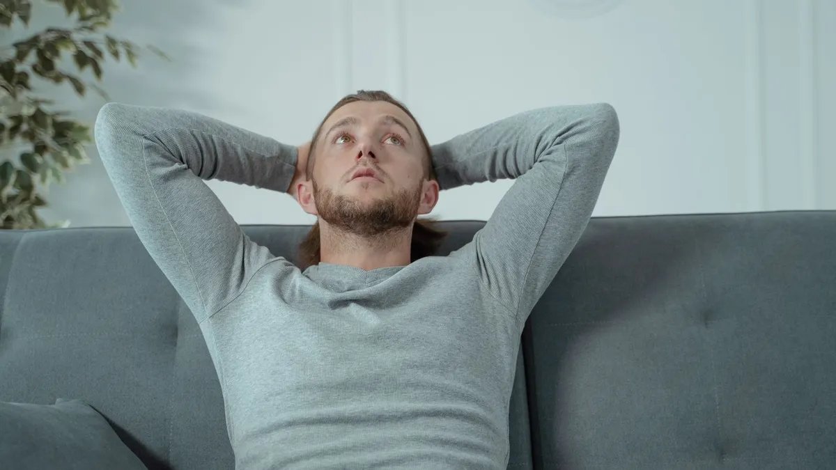 Mann lehnt sich auf dem Sofa zurück, die Hände im Nacken, und blickt gedankenverloren nach oben
