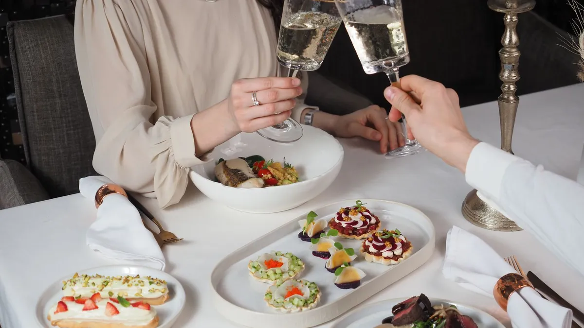 Zwei Personen stoßen an einem weiß gedeckten Restauranttisch mit Sekt an, davor kleine Gerichte