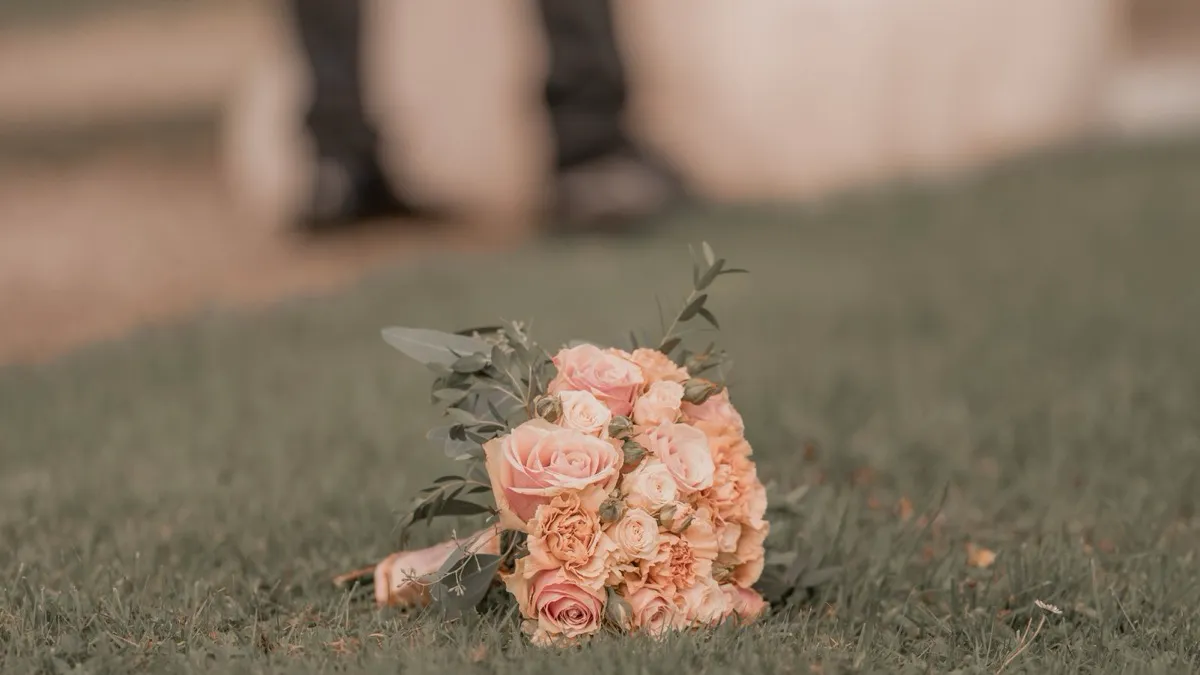 Brautstrauß aus rosafarbenen Rosen liegt auf einer Wiese, im unscharfen Hintergrund steht ein Paar