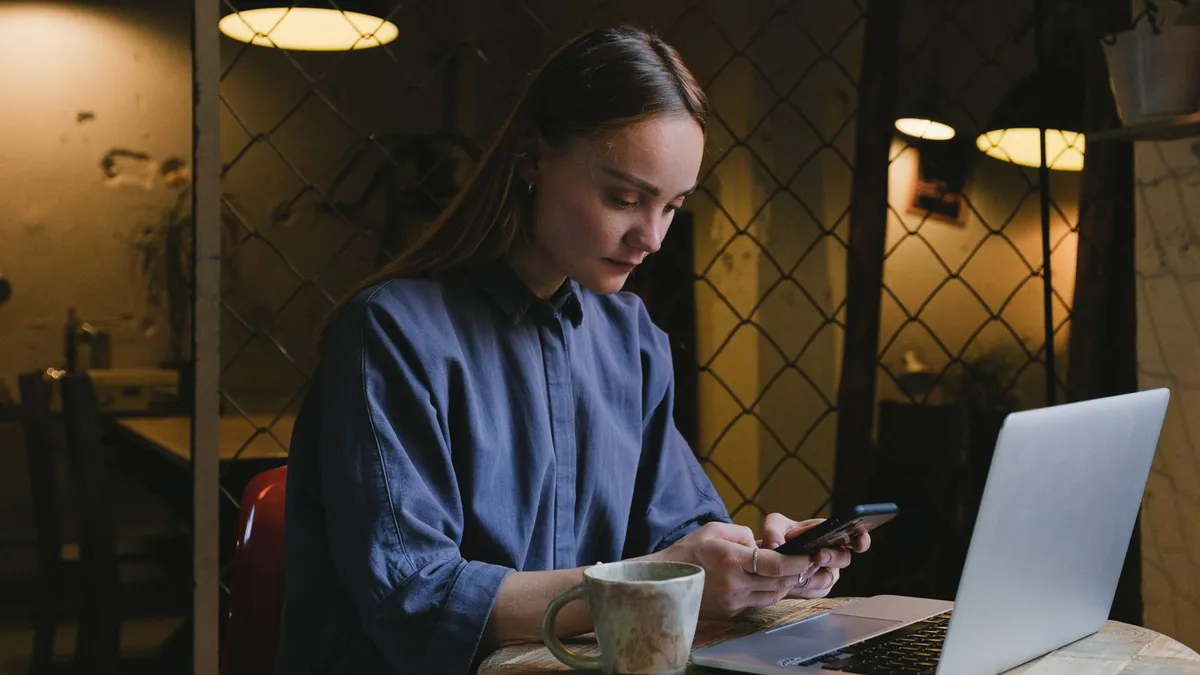 Konzentrierte Frau sitzt in einem Café vor ihrem Laptop und tippt auf ihrem Smartphone