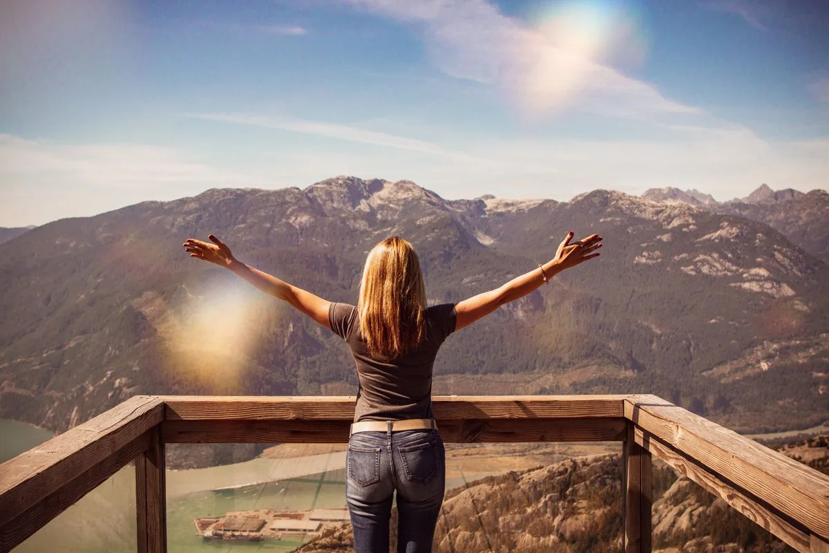 Frau steht mit weit ausgebreiteten Armen auf einer Aussichtsplattform vor einer Bergkulisse