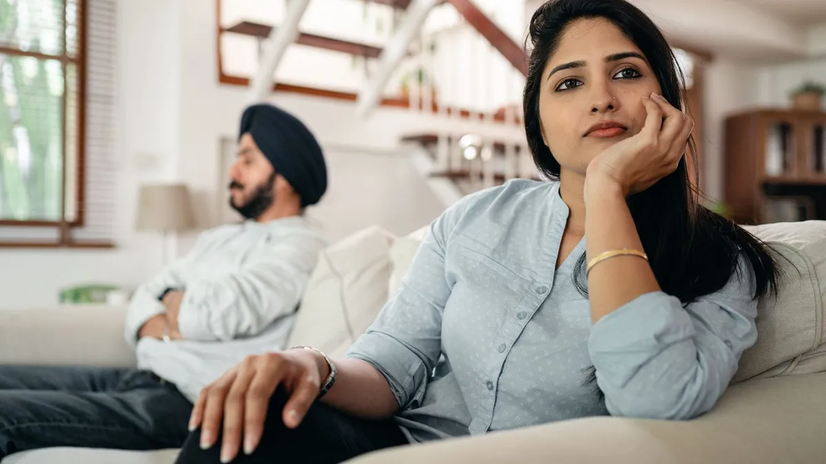 Eine Frau stützt auf dem Sofa nachdenklich den Kopf in die Hand, neben ihr lehnt ihr Partner abgewandt