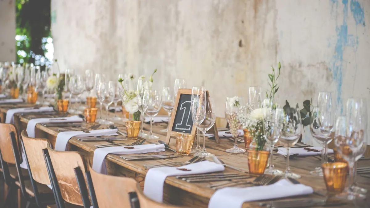 Lange Hochzeitstafel aus Holz mit Weingläsern, weißen Servietten, kleinen Blumenvasen und einer Kreidetafel mit der Tischnummer 1