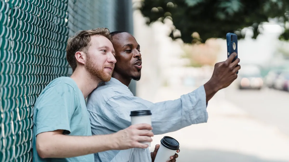 Zwei junge Männer machen lachend ein Selfie mit dem Smartphone auf der Straße, in der Hand Kaffeebecher