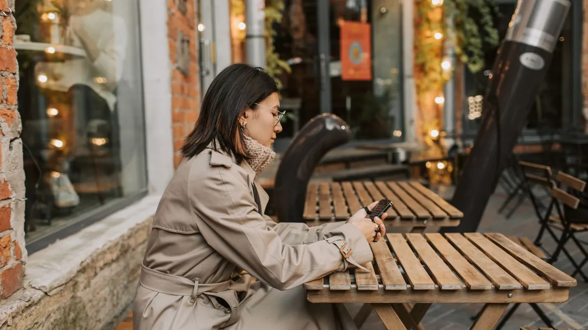 Frau im Trenchcoat sitzt an einem Holztisch vor einem Café und schaut auf ihr Smartphone