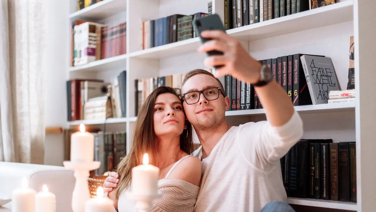 Paar macht ein Selfie vor einem Bücherregal, im Vordergrund stehen brennende Kerzen