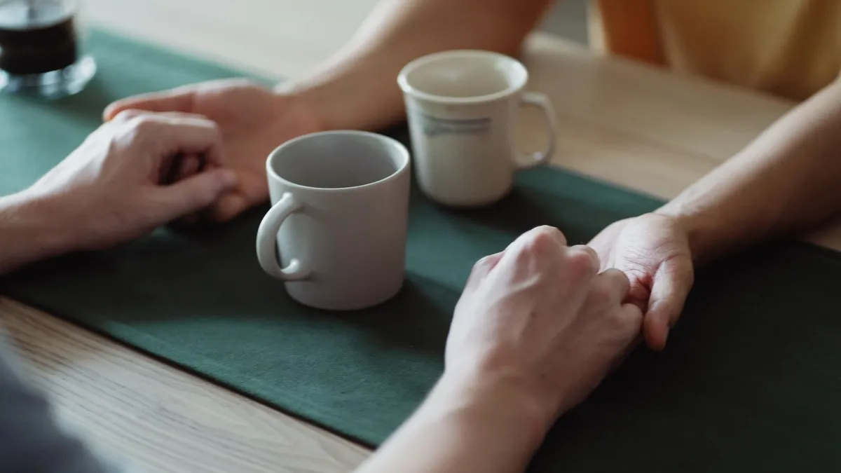 Zwei Paar Hände halten sich über einem Tisch mit zwei Kaffeetassen, Gesichter sind nicht zu sehen