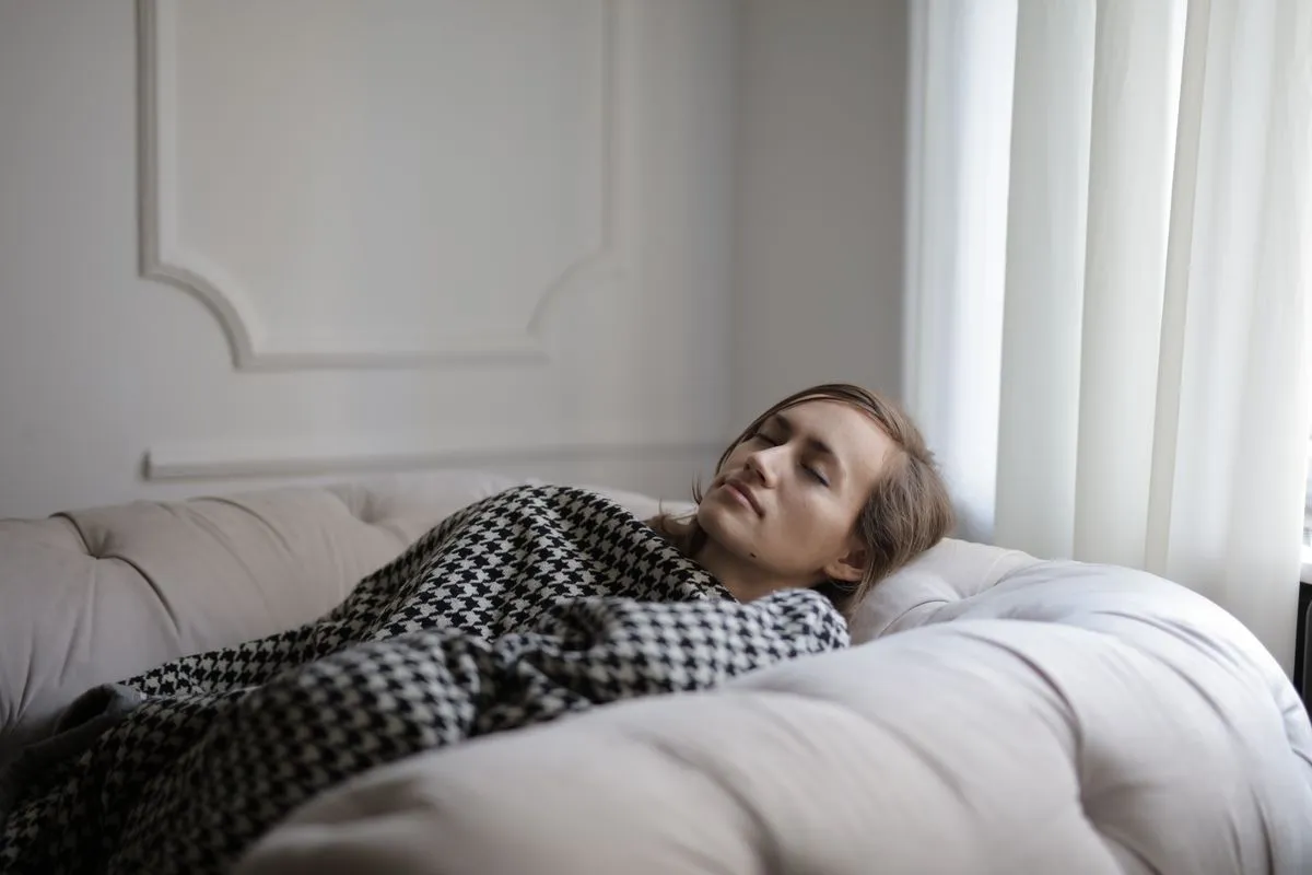 Junge Frau ruht mit geschlossenen Augen unter einer Decke auf dem Sofa und lädt ihre Energie auf