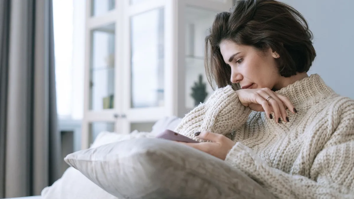 Nachdenkliche Frau in cremefarbenem Strickpullover sitzt mit einem Kissen auf dem Sofa und blickt auf ihr Handy
