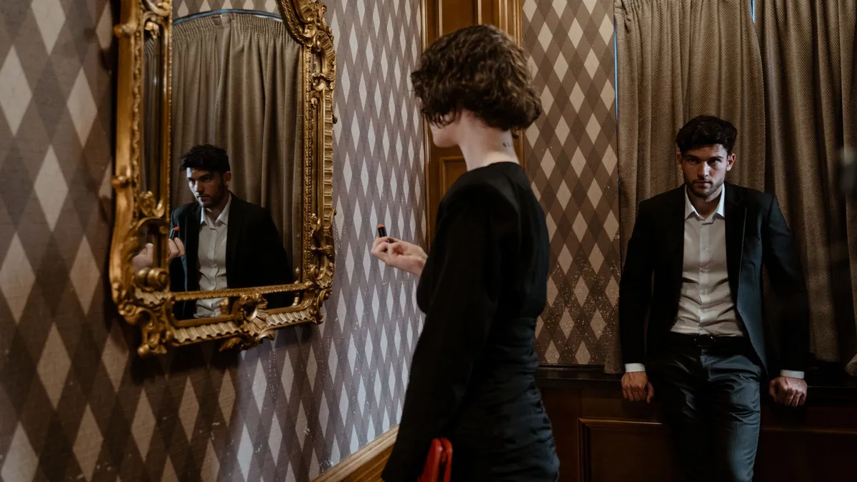 Elegant gekleidetes Paar in einem Raum mit Goldspiegel, sie schaut in den Spiegel, er lehnt an der Wand