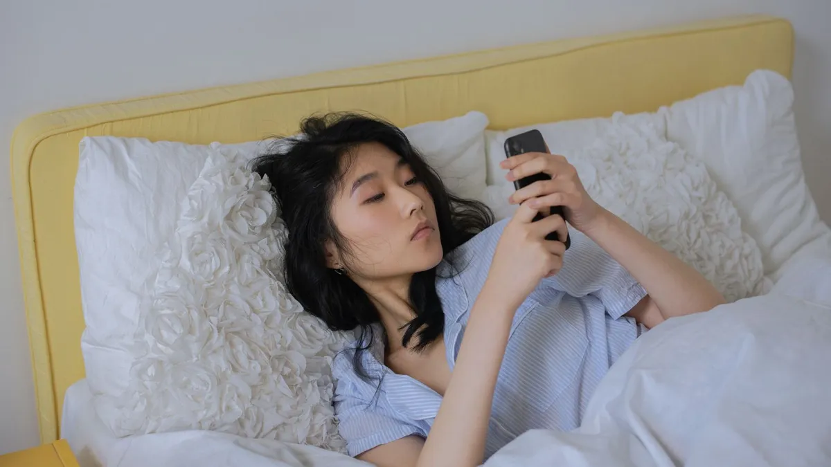 Frau liegt im Bett und blickt nachdenklich auf ihr Smartphone
