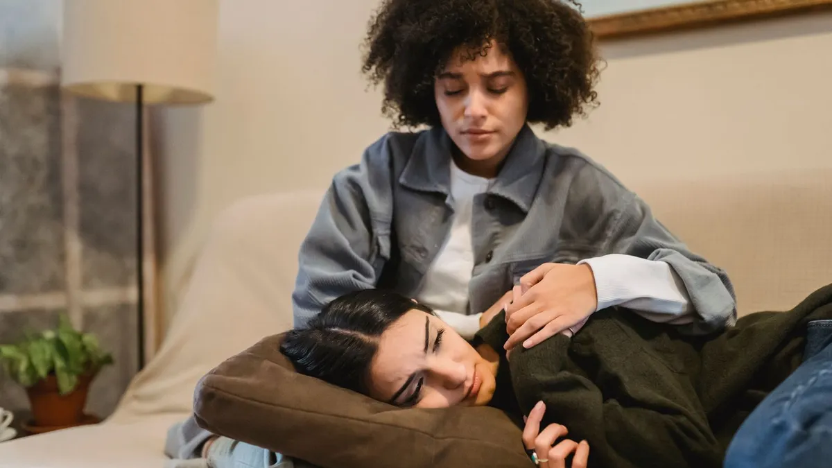 Eine Frau mit lockigem Haar tröstet auf dem Sofa ihre Freundin, die erschöpft den Kopf in ihren Schoß legt