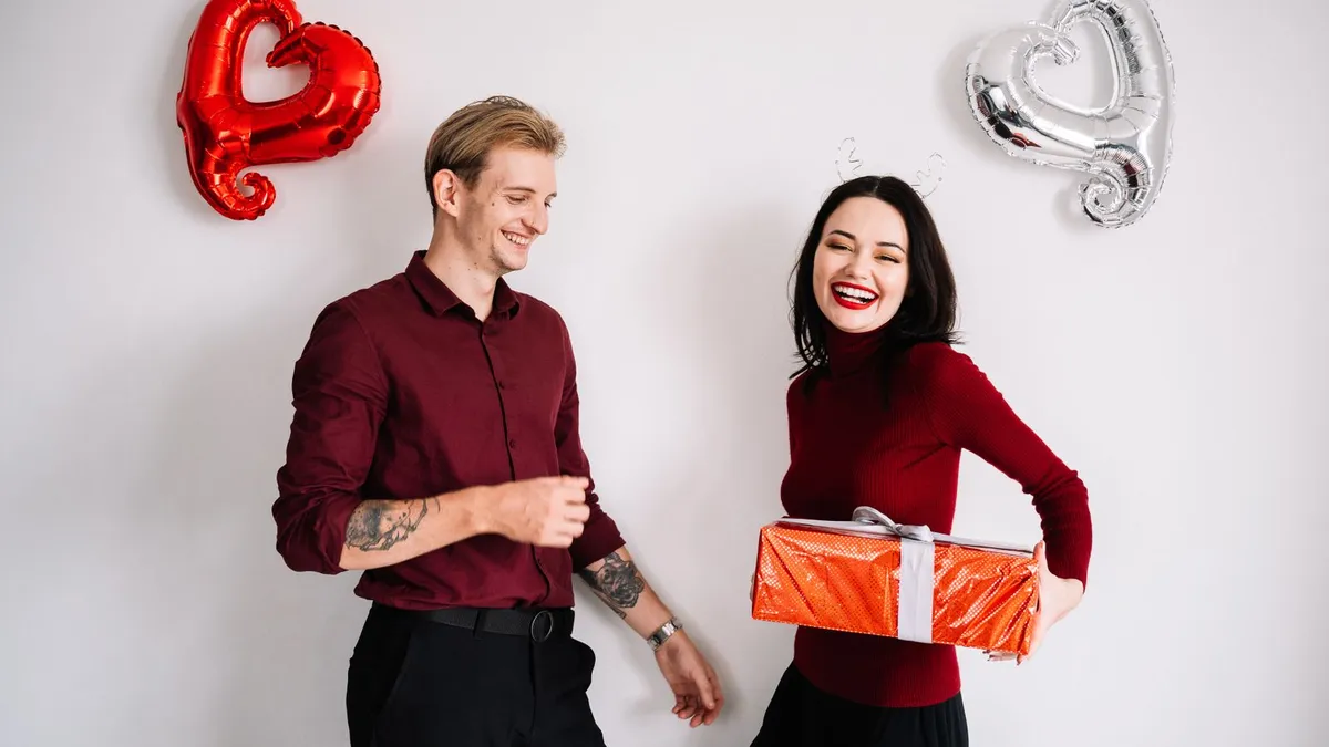 Lachendes Paar vor einer Wand mit Herzluftballons, die Frau hält ein rotes Geschenk